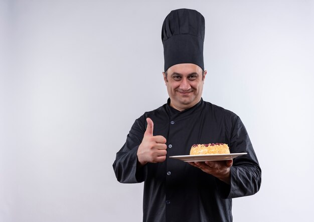 Pleased middle-aged male cook in chef uniform holding cake on plate his thumb up on isolated white wall with copy space