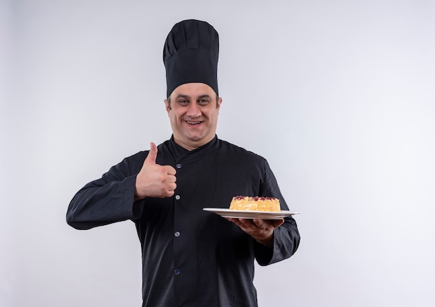 Довольный мужчина средних лет повар в униформе шеф-повара держит торт на тарелке большим пальцем на изолированной белой стене с копией пространства