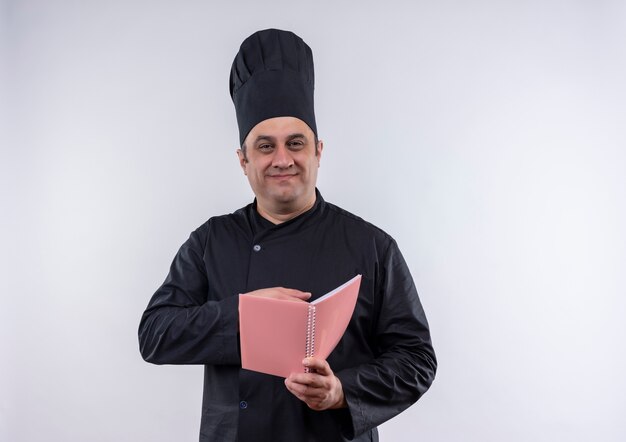 Pleased middle-aged male cook in chef uniform holding notebook on isolated white wall with copy place