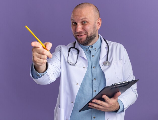 Pleased middle-aged male doctor wearing medical robe and stethoscope holding clipboard and pencil looking and pointing at camera 