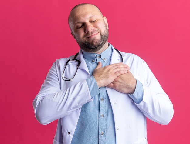 Felice medico maschio di mezza età che indossa tunica medica e stetoscopio tenendo la mano sul petto con gli occhi chiusi isolati su parete rosa on