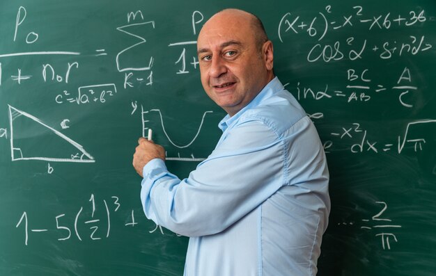 pleased middle-aged male teacher standing in front blackboard holding stranded for board