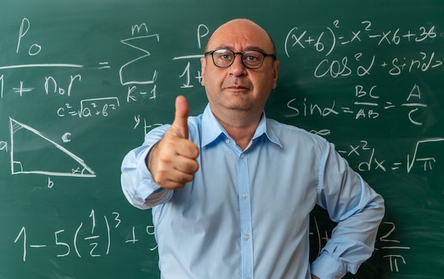 pleased middle-aged male teacher wearing glasses standing in front blackboard showing thumb up