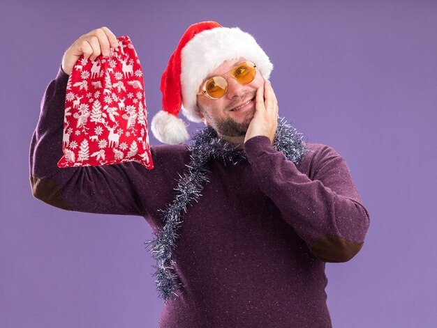 pleased middle-aged man wearing santa hat and tinsel garland around neck with glasses holding christmas gift sack looking at side keeping hand on face isolated on purple background