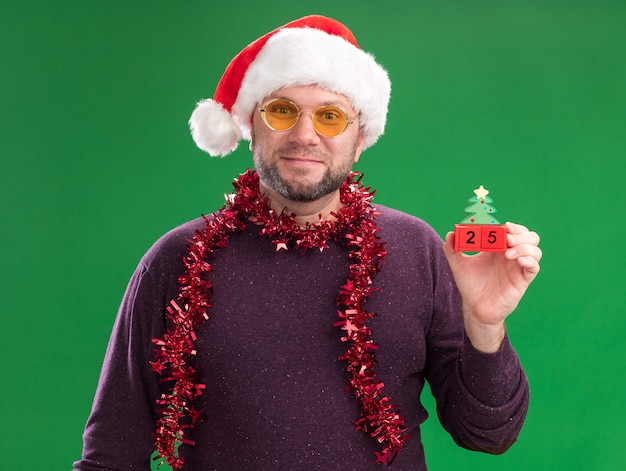 Довольный мужчина средних лет в новогодней шапке и мишурной гирлянде на шее в очках, держащий елочную игрушку с датой на зеленой стене
