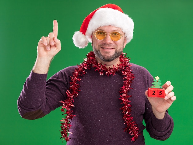 Довольный мужчина средних лет в новогодней шапке и мишурной гирлянде на шее в очках, держащий елочную игрушку с датой