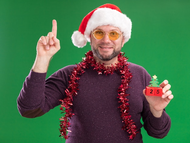 Pleased middle-aged man wearing santa hat and tinsel garland around neck with glasses holding christmas tree toy with date