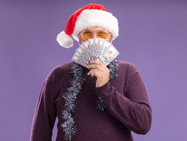 보라색 벽에 고립 된 뒤에서 돈을 들고 안경 목에 산타 모자와 반짝이 갈 랜드를 입고 기쁘게 중년 남자