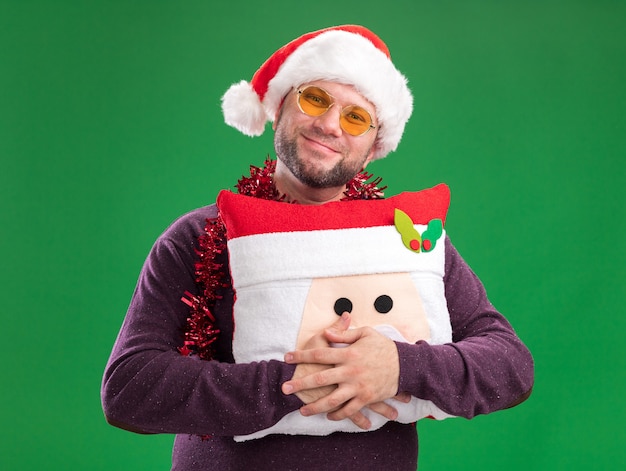 Pleased middle-aged man wearing santa hat and tinsel garland around neck with glasses holding santa claus pillow  isolated on green wall