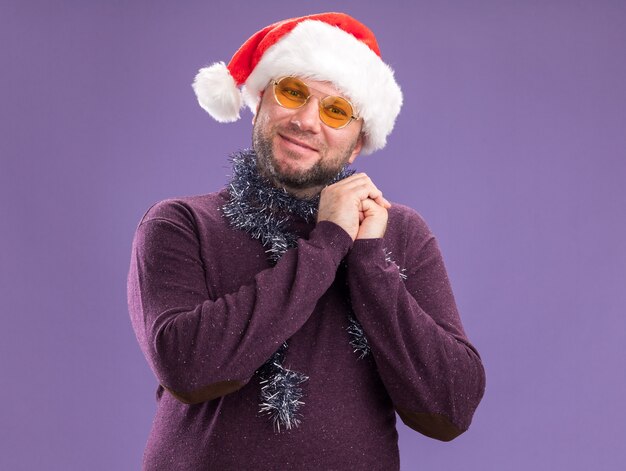 Pleased middle-aged man wearing santa hat and tinsel garland around neck with glasses  keeping hands together isolated on purple wall