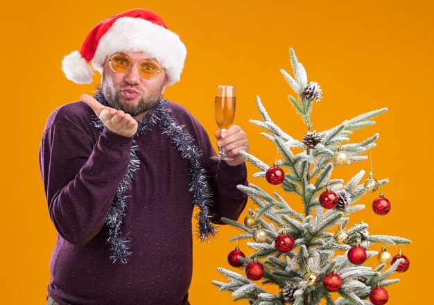 Pleased middle-aged man wearing santa hat and tinsel garland around neck with glasses standing near decorated christmas tree holding glass of champagne 