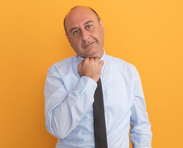 Pleased middle-aged man wearing white t-shirt with tie holding collar isolated on orange wall
