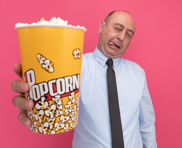 Pleased middle-aged man wearing white t-shirt with tie holding out bucket of popcorn at camera isolated on pink wall