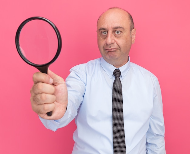 Pleased middle-aged man wearing white t-shirt with tie holding out magnifier  isolated on pink wall