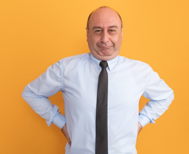 Pleased middle-aged man wearing white t-shirt with tie putting hands on waist isolated on orange wall