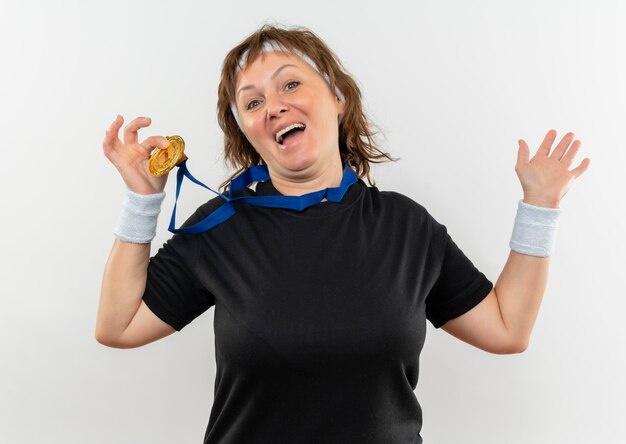 Pleased middle aged sporty woman in black t-shirt with headband and gold medal around her neck showing it smiling cheerfully standing over white wall