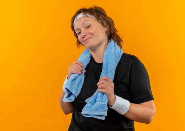 Pleased middle aged sporty woman in black t-shirt with headband and with towel on shoulder with confident smile on face standing over orange wall