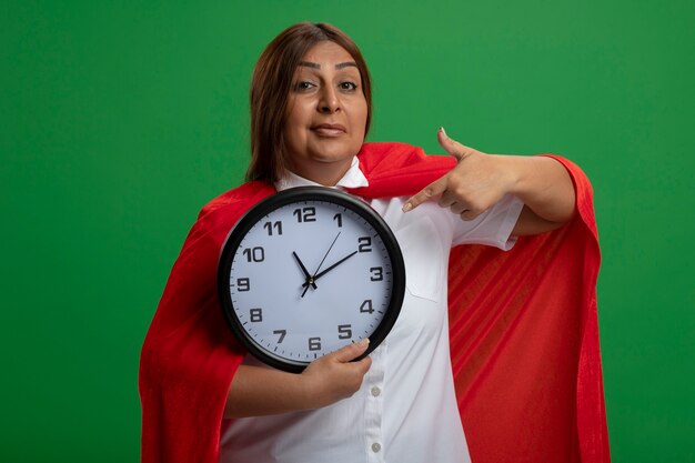 Pleased middle-aged superhero female holding and points at wall clock isolated on green background