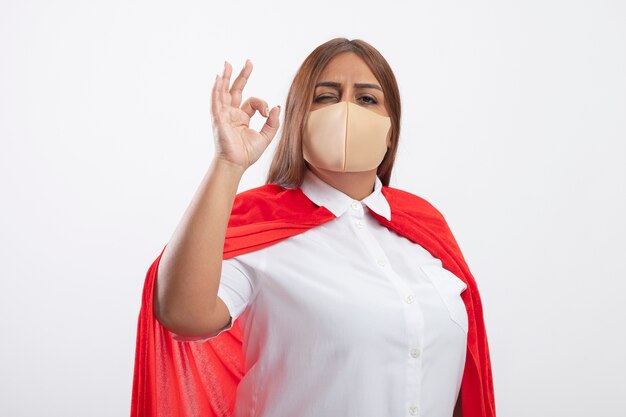 Довольная женщина-супергерой средних лет в медицинской маске показывает нормальный жест, изолированный на белом
