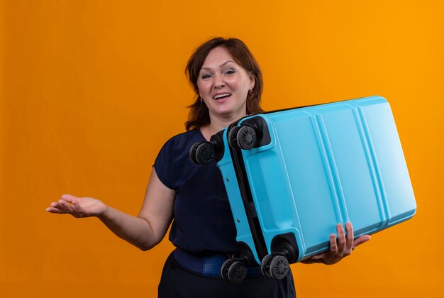 Довольная женщина-путешественница средних лет держит чемодан и показывает рукой из стороны в сторону на изолированном оранжевом фоне