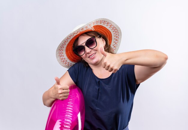 Pleased middle-aged traveler woman wearing glasses with hat holding inflatable ring her thumbs up on isolated white wall