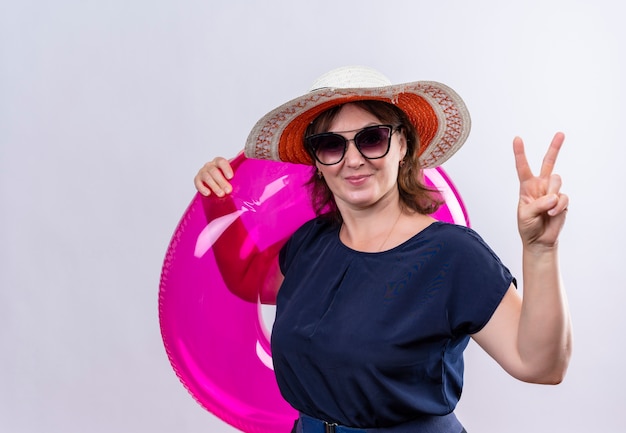 Pleased middle-aged traveler woman wearing glasses with hat holding inflatable ring showing peace gesture on isolated white wall