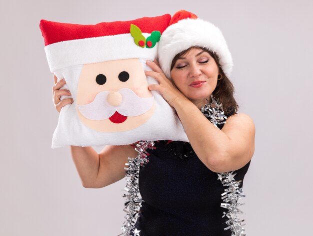 Pleased middle-aged woman wearing santa hat and tinsel garland around neck holding santa claus pillow touching head with it with closed eyes isolated on white background