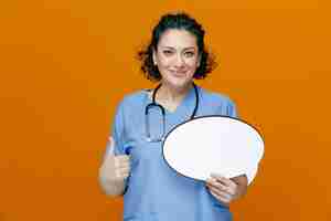 Free photo pleased middleaged female doctor wearing uniform and stethoscope around her neck looking at camera showing chat bubble and thumb up isolated on orange background