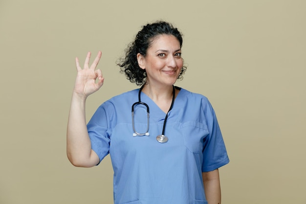 Pleased middleaged female doctor wearing uniform and stethoscope around neck looking at camera showing ok sign isolated on olive background