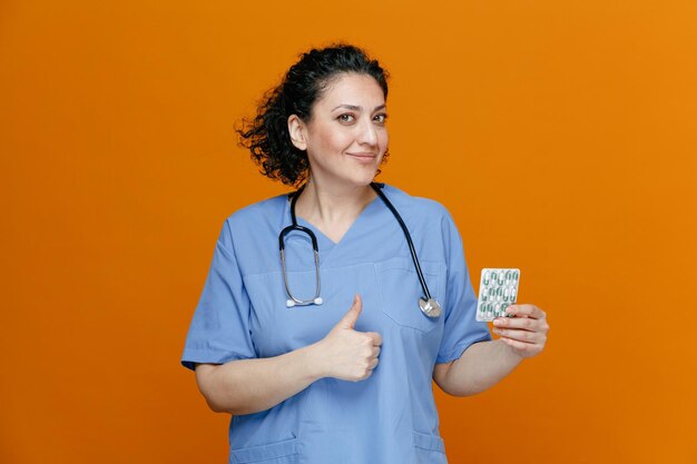 pleased middleaged female doctor wearing uniform and stethoscope around neck showing pack of pills and thumb up looking at camera isolated on orange background