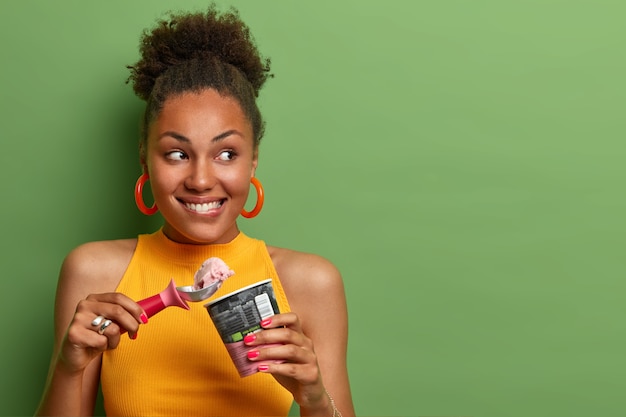 Foto gratuita piacevole ragazza millenaria con i capelli ricci, morde le labbra e mangia un delizioso gelato con appetito, gode di un sapore naturale, brividi durante il giorno d'estate, guarda volentieri da parte, spazio vuoto sul muro verde