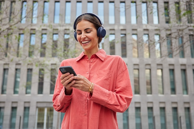 La ragazza millenaria soddisfatta con i capelli scuri si gode il tempo libero ascolta la playlist musicale utilizza le moderne cuffie wireless per smartphone ha la città che cammina vestita con una camicia rossa posa contro un edificio moderno