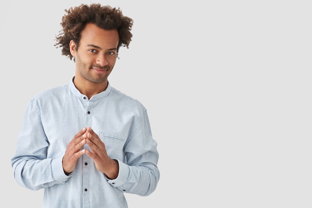 Free photo pleased mixed race young man keeps hands together, has intriguing expression and intention to do something, going to share his plans with friend, isolated over white blank wall