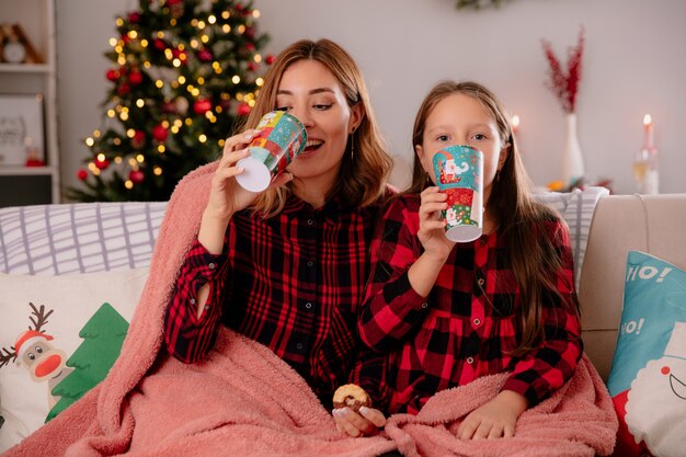 紙コップとクッキーを持ち、毛布で覆われたソファに座り、家でクリスマスを楽しむ母と娘