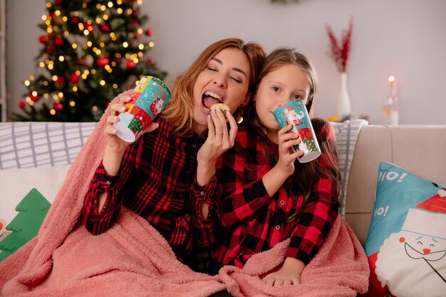 pleased mother and daughter holding paper cups and eating biscuits sitting on couch covered with blanket and enjoying christmas time at home