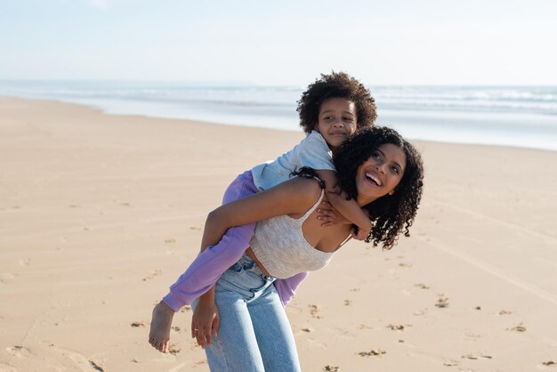 행복한 엄마와 딸이 해변에서 시간을 보내고 있습니다. 아프리카계 미국인 가족은 걷고, 웃고, 놀고, 등에 타고 있습니다. 여가, 가족 시간, 부모 개념