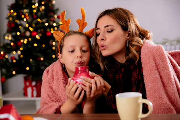 довольные мать и дочь с одеялом держат и задувают свечу, сидя за столом, наслаждаясь Рождеством дома