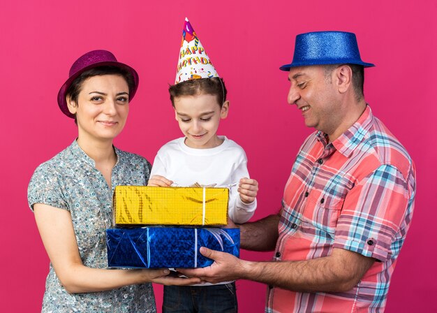 pleased mother and father with party hats holding gift boxes together standing with their son isolated on pink wall with copy space