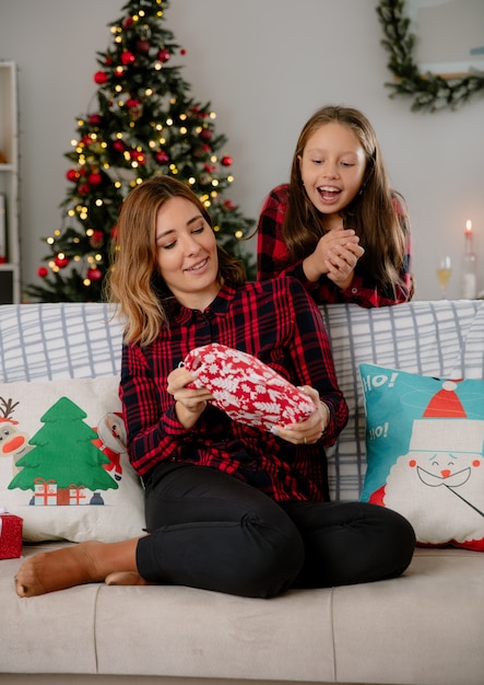 pleased mother holds and excited daughter looks at gift package sitting on couch and enjoying christmas time at home