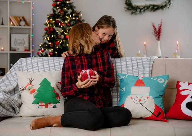 pleased mother holds gift box and kisses daughter sitting on couch and enjoying christmas time at home