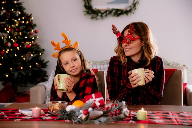 家でクリスマスの時間を楽しんでいるテーブルに座ってカップを保持している娘を見て、トナカイの眼鏡をかけた母が喜んでいる