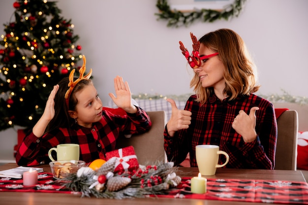 家でクリスマスの時間を楽しんでいるテーブルに座っている娘を見て、トナカイの眼鏡をかけた母が喜んでいる