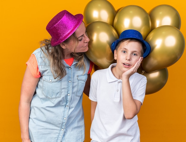 pleased mother wearing purple party hat and holding helium balloons looking at her surprised son with blue party hat putting hand on face isolated on orange wall with copy space