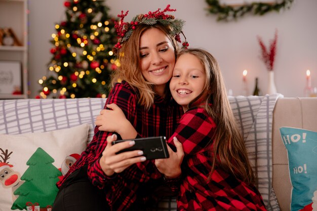 ヒイラギの花輪を持つ母と娘がソファに座って電話で何かを見て、家でクリスマスの時間を楽しんでいる