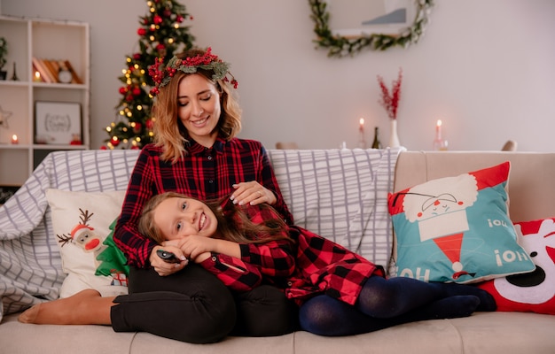 Pleased mother with holly wreath holds tv remote and looks at daughter lying on her lap sitting on couch and enjoying christmas time at home