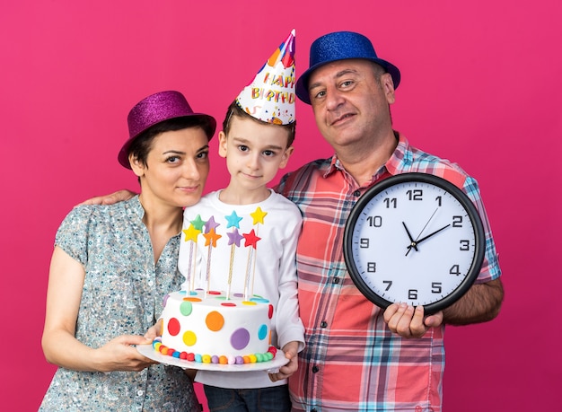 誕生日ケーキを保持している紫色のパーティハットとコピースペースでピンクの壁に隔離された息子と一緒に立っている時計を保持している青いパーティハットで笑顔の父を喜んで母親