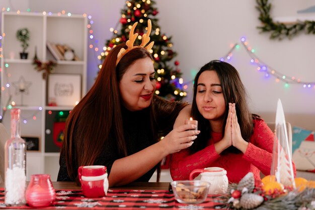トナカイのカチューシャを持って喜んでいる母親は、家でクリスマスの時間を楽しんでいるテーブルに座っている娘と一緒にキャンドルを持って見ています