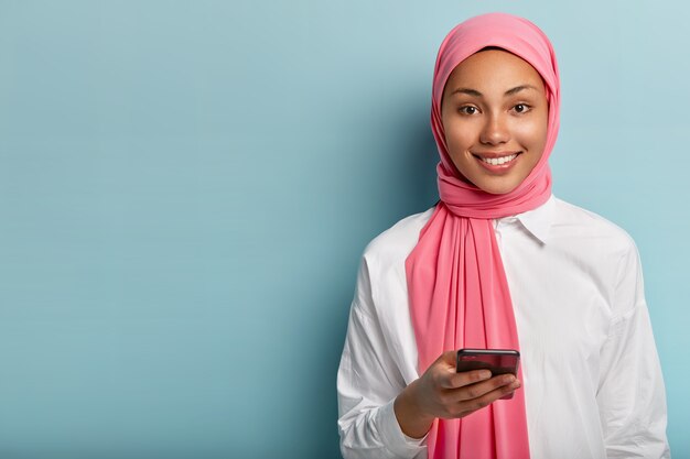 Pleased Muslim woman uses cell phone to socialize, gives reply in online chat, posts something in social networks