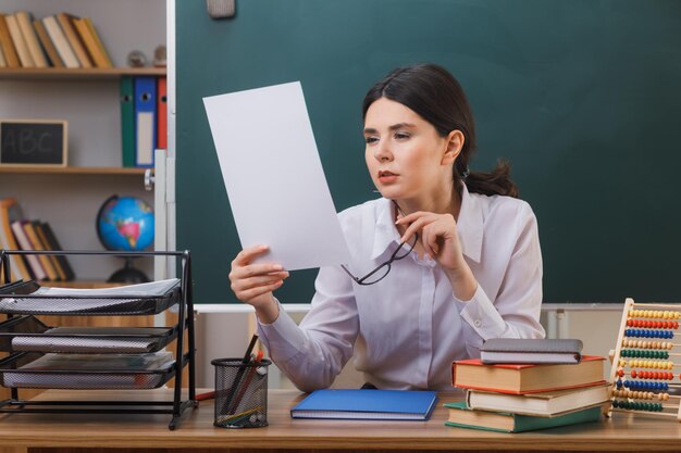 pleased ng female teacher holding glasses and looking at paper in her hand sitting at desk with school tools in classroom