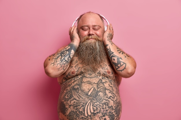 Pleased obese fat man enjoys listening favorite music in stereo headphones, poses with naked belly, has tattooed arms and tummy, overweight because of eating fast food, isolated on pink wall
